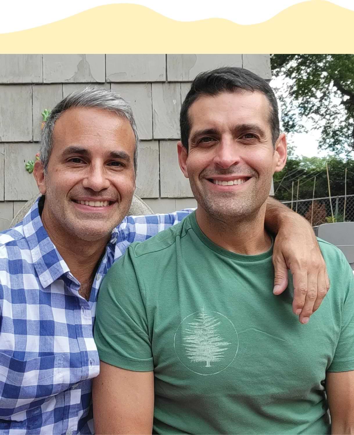 Dr. Omar Farias photographed with his husband standing outside a house, both smile with his husband arms around the doctors shoulder