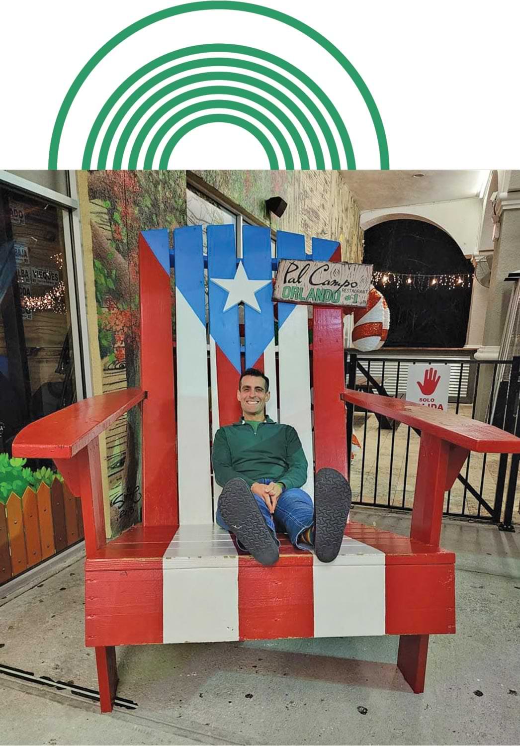 Dr. Omar Farias smiles while sitting back in a oversized wooden adirondack chair painted with the Texas flag and holdig a sign the reads "Pal Campo Restaurant Orlando #1"
