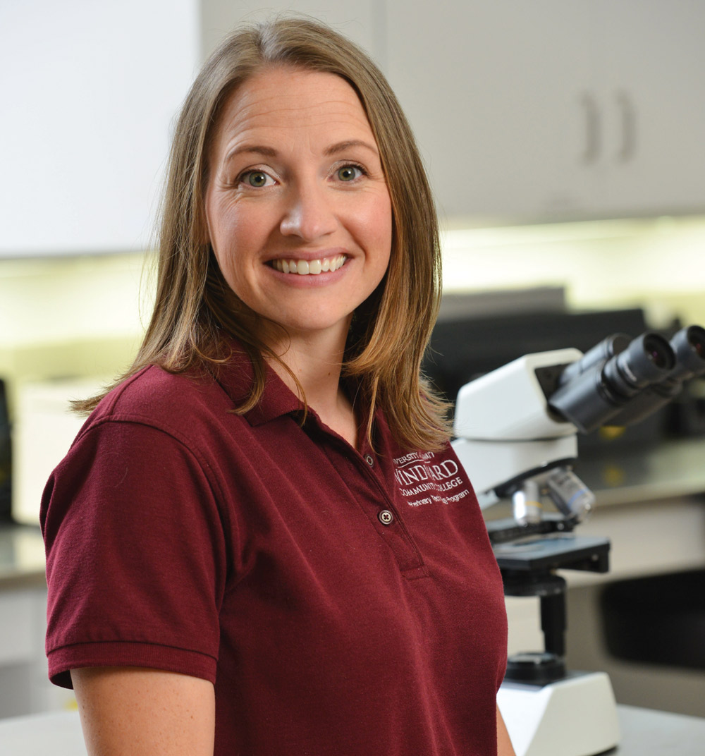 A smiling woman in a maroon polo shirt stands in a laboratory next to a microscope.