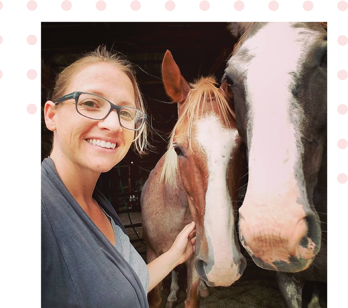 A person with glasses smiles next to two horses in a barn.