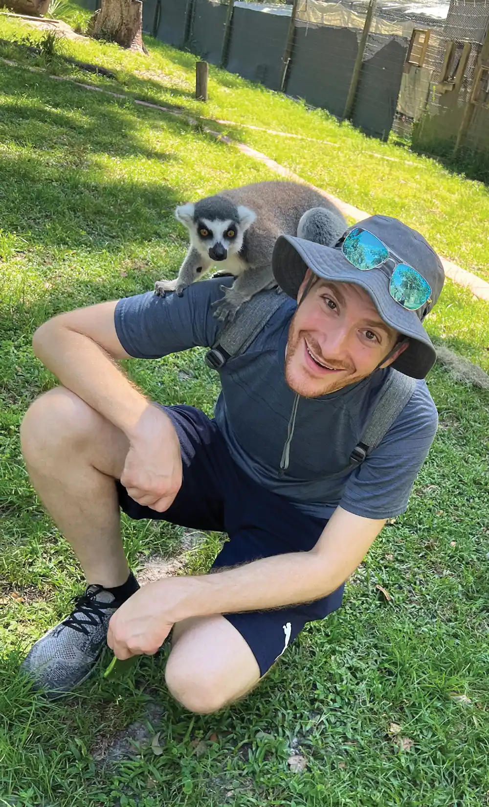A person crouching on grass with a lemur on their shoulder, wearing a gray hat and reflective sunglasses.
