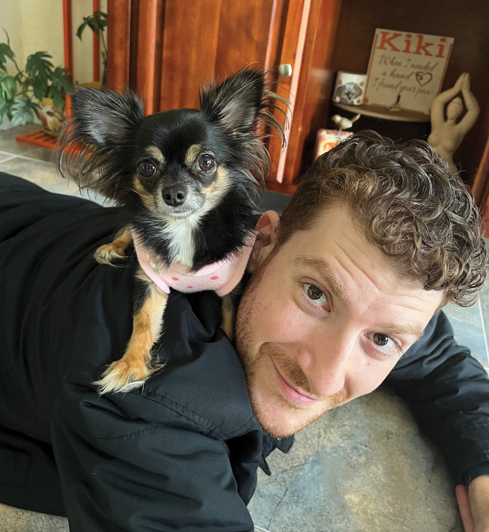 Small dog sitting on a person who is lying on a tiled floor, with a wooden cabinet in the background.